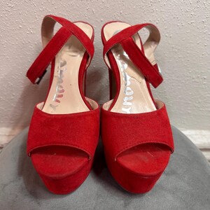 May include: Pair of red platform heels with a peep-toe design. The heels have a thick platform sole and a criss-cross ankle strap. The word "Amour" is printed on the insole. The shoes are made of a suede-like material.
