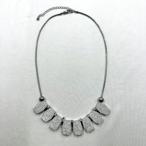 May include: A silver necklace featuring a series of textured, rectangular pendants. The necklace has a fine chain and a lobster clasp. The pendants are arranged in a curved row, creating a statement piece. The necklace is displayed on a white surface.