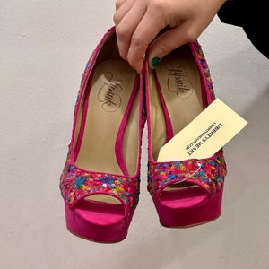 Puede incluir: Un par de zapatos de tacón alto rosa con punta abierta y un diseño de lentejuelas de colores. Los zapatos están etiquetados como "Lipstik" y "Liberty's Heart".