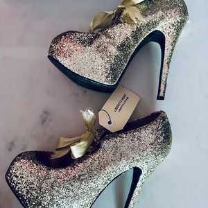 May include: A pair of gold glitter platform heels with a satin ribbon bow. The heels are labelled "Liberty Heart" and "Liberty London".