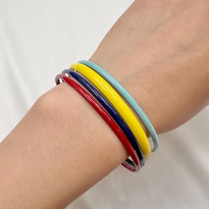 May include: A set of four thin, smooth bangle bracelets in solid colours. The stack includes one red, one dark blue, one bright yellow, and one light blue bangle, all featuring a glossy finish and a simple, rounded, circular design.