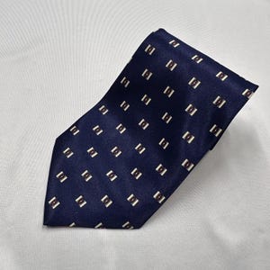 Puede incluir: Una corbata azul marino con un patrón geométrico repetitivo de pequeños rectángulos blancos con puntos de color rojo oscuro en el centro. La corbata tiene un acabado liso y ligeramente reflectante con un extremo puntiagudo clásico.