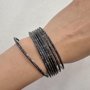 Pode incluir: Um conjunto de pulseiras finas de cor escura com uma superfície texturizada e partículas de glitter douradas incrustadas. As pulseiras estão dispostas em uma pilha, com uma pulseira separada do grupo principal, mostrando um perfil circular fino.