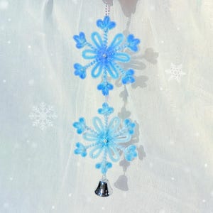 May include: A decorative hanging ornament featuring two blue snowflake designs connected by a string. The snowflakes are made of fluffy material and adorned with small beads. A silver bell hangs at the bottom, adding a festive touch.