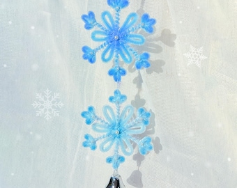 Op de afbeelding: Een decoratief hangornament met twee blauwe sneeuwvlokontwerpen die met een touwtje zijn verbonden. De sneeuwvlokken zijn gemaakt van zacht materiaal en versierd met kleine kralen. Een zilveren bel hangt aan de onderkant.