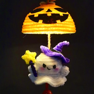 May include: A handmade Halloween decoration featuring a smiling ghost wearing a purple witch hat and holding a yellow star wand. The ghost hangs below a glowing orange pumpkin with a carved face. A red bead hangs below the ghost.