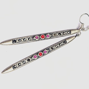 Puede incluir: Un par de pendientes largos y delgados de metal en tono plateado antiguo con forma de aguja puntiaguda. Cada pendiente está decorado con una fila de pequeños cristales negros y un grupo central de un cristal rojo y dos rosas.