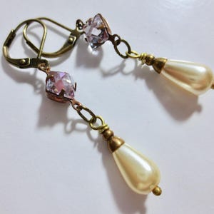 May include: A pair of vintage-style earrings. Each earring features a faceted pink crystal, connected by a chain to a teardrop-shaped faux pearl. The earrings have an antique brass finish and lever-back closures.