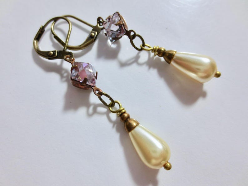 May include: A pair of vintage-style earrings. Each earring features a faceted pink crystal, connected by a chain to a teardrop-shaped faux pearl. The earrings have an antique brass finish and lever-back closures.