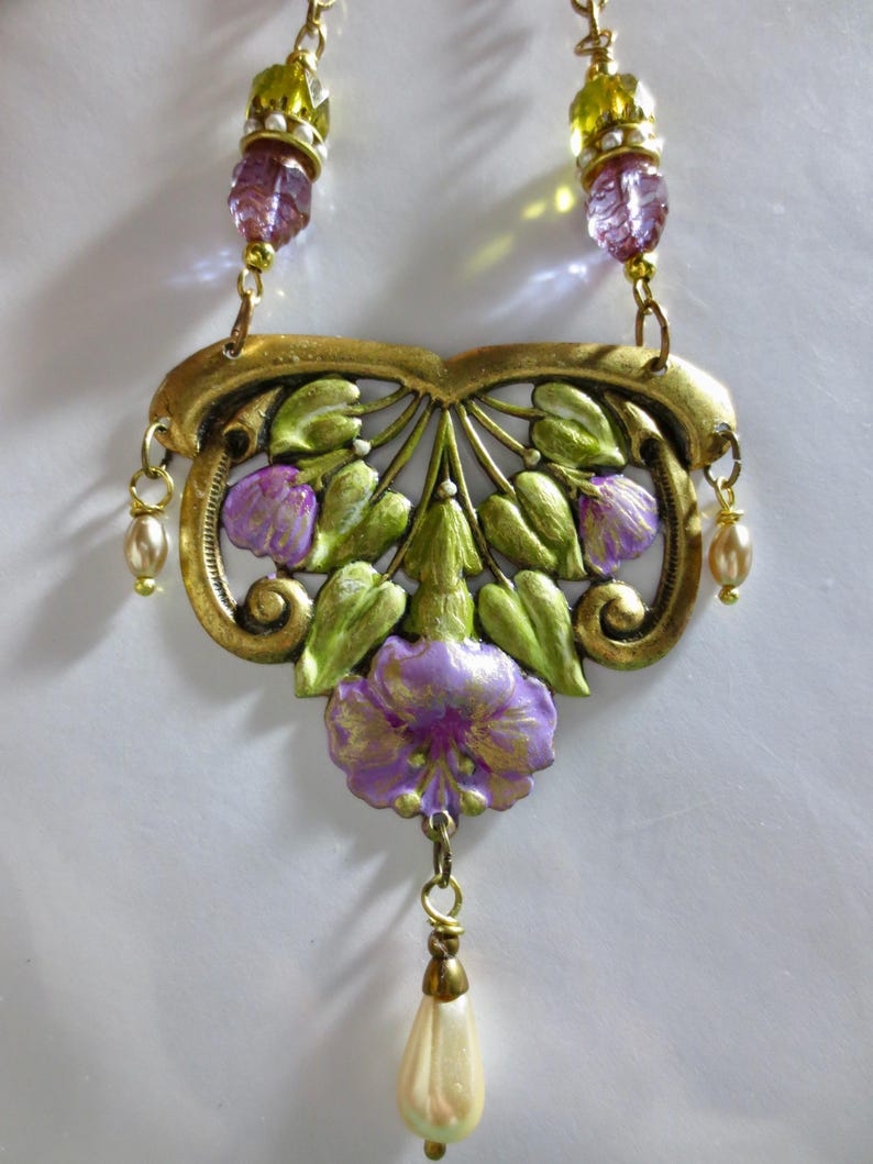 May include: A gold-toned necklace with a floral pendant. The pendant features painted purple flowers and green leaves. The necklace has a pearl drop and is adorned with purple and green beads, evoking a vintage aesthetic.
