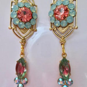 Op de afbeelding: Een paar goudkleurige oorbellen met een Art Deco-ontwerp. Elke oorbel heeft een centrale perzikkleurige kristal omringd door lichtblauwe kristallen. Een groene en rode kristal hangt eronder, met drie kleinere blauwe kristallen.