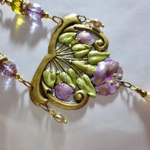 May include: A vintage-style necklace with a floral pendant. The pendant features a gold-toned metal frame with green leaves and purple flowers. The necklace has beaded strands with yellow and purple beads, and a cream-colored teardrop charm.