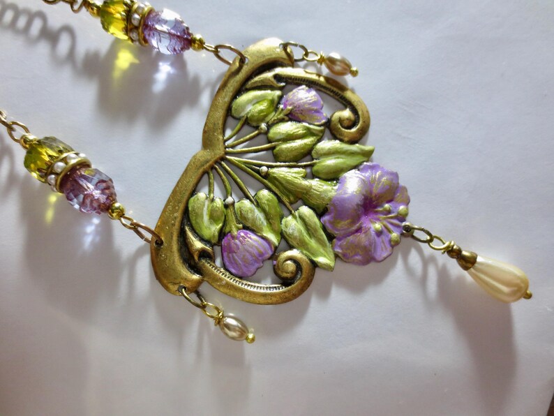 May include: A vintage-style necklace with a floral pendant. The pendant features a gold-toned metal frame with green leaves and purple flowers. The necklace has beaded strands with yellow and purple beads, and a cream-colored teardrop charm.