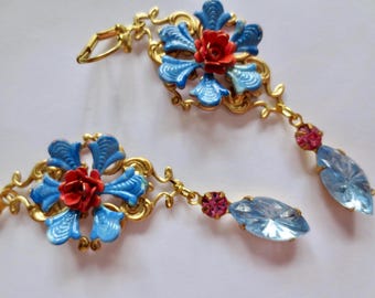 May include: A pair of gold-tone earrings featuring a central floral motif with blue and red enamel petals. Each earring includes a small pink crystal accent and a dangling, faceted, light blue marquise-shaped crystal drop.