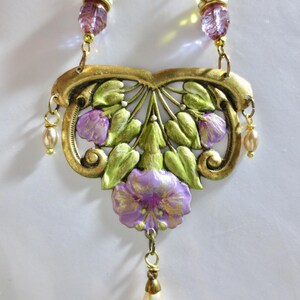 May include: A vintage-style necklace with a floral design. The pendant is gold-toned with green leaves and purple flowers. The necklace has a drop pendant and is accented with faux pearls and faceted beads.