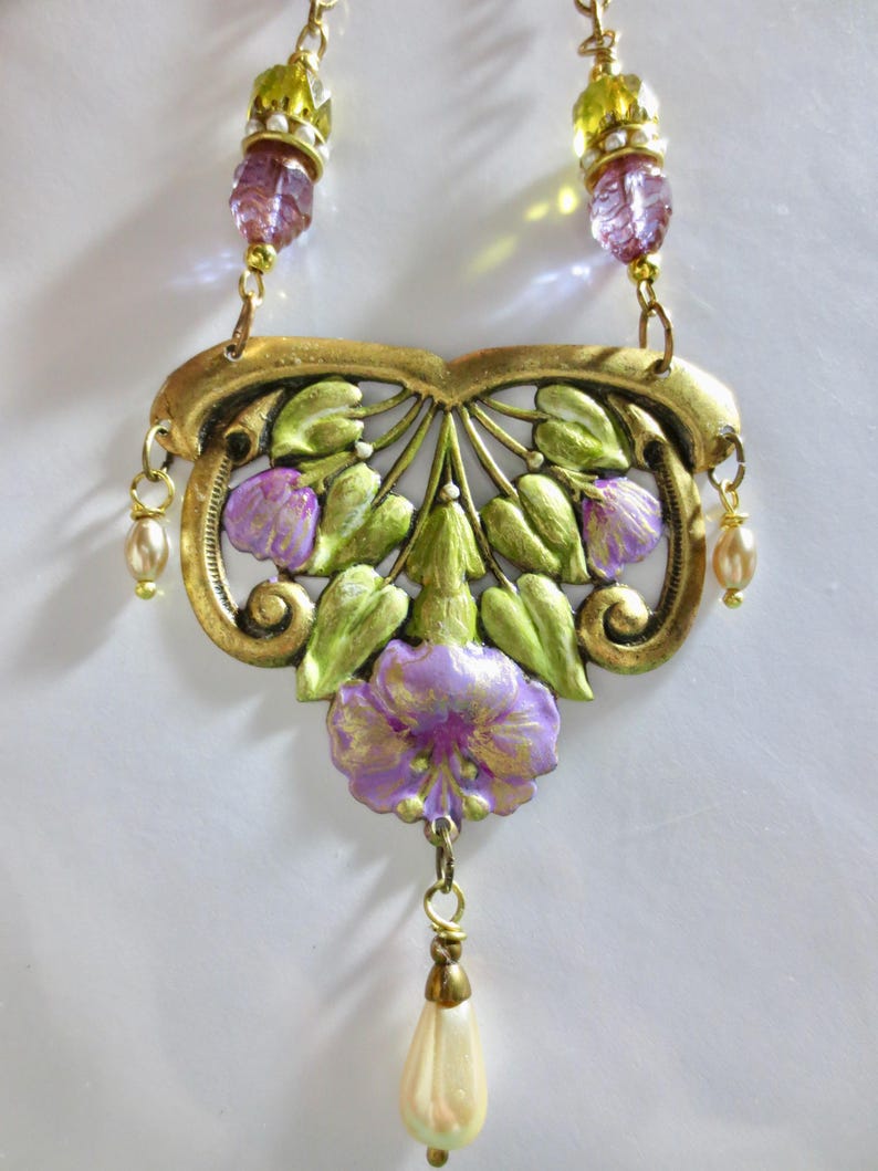 May include: A vintage-style necklace with a floral design. The pendant is gold-toned with green leaves and purple flowers. The necklace has a drop pendant and is accented with faux pearls and faceted beads.