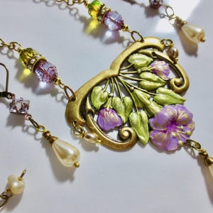 May include: A vintage-style necklace and earring set. The necklace features a gold-toned pendant with purple flowers and green leaves. The earrings and necklace have pearl and crystal accents. The jewelry is set against a white background.