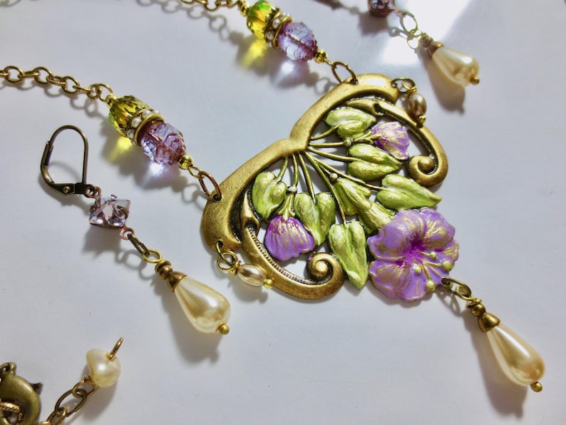 May include: A vintage-style necklace and earring set. The necklace features a gold-toned pendant with purple flowers and green leaves. The earrings and necklace have pearl and crystal accents. The jewelry is set against a white background.