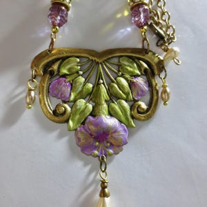 May include: A vintage-style necklace with a gold-tone metal filigree pendant featuring purple flowers and green leaves. The necklace has a delicate chain with purple and green beads, and a pear-shaped faux pearl drop. The overall design is ornate and detailed.