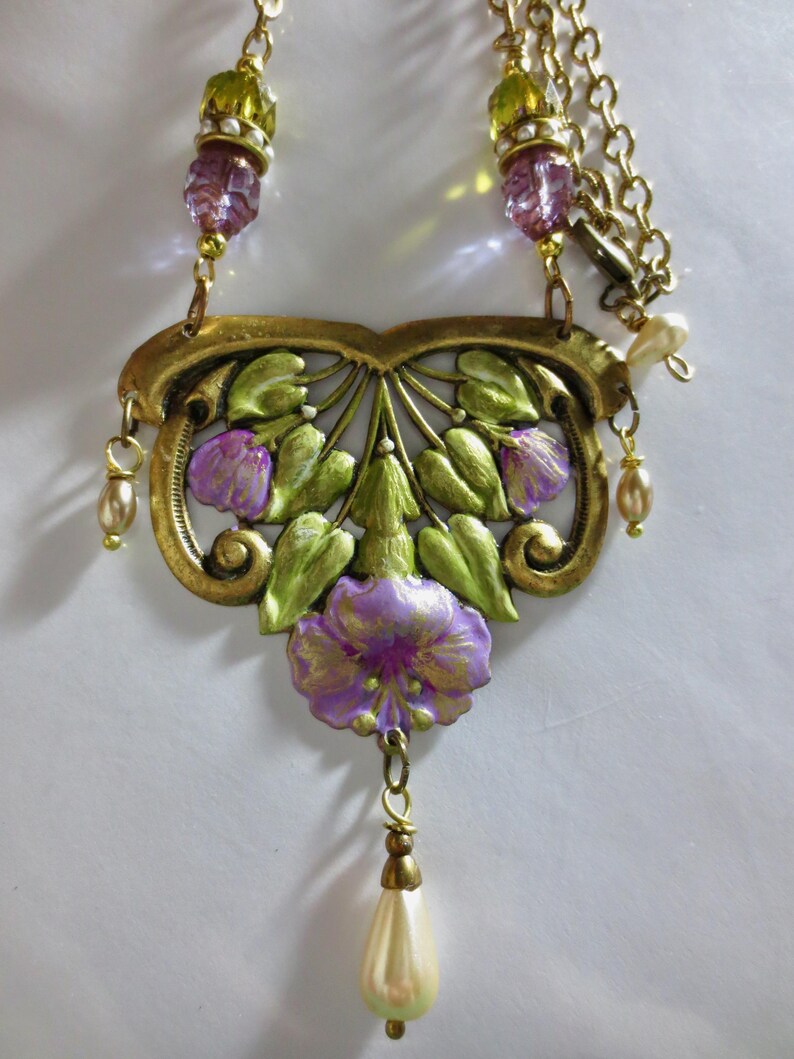 May include: A vintage-style necklace with a gold-tone metal filigree pendant featuring purple flowers and green leaves. The necklace has a delicate chain with purple and green beads, and a pear-shaped faux pearl drop. The overall design is ornate and detailed.