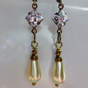 May include: A pair of vintage-style dangle earrings. Each earring features a clear, square-cut crystal at the top, connected to a gold-toned wire. Below the crystal hangs a pear-shaped, cream-colored faux pearl. The earrings have a classic, elegant design.