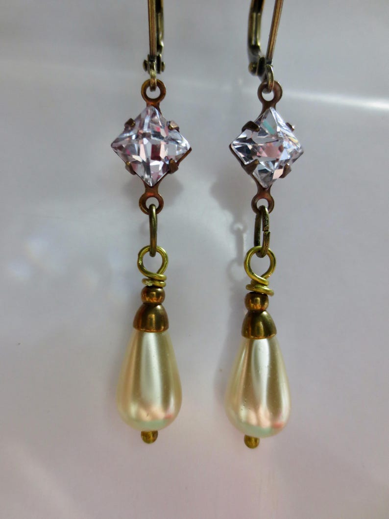 May include: A pair of vintage-style dangle earrings. Each earring features a clear, square-cut crystal at the top, connected to a gold-toned wire. Below the crystal hangs a pear-shaped, cream-colored faux pearl. The earrings have a classic, elegant design.