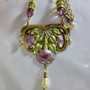 May include: A vintage-style necklace featuring a floral pendant with purple and green painted details. The pendant is suspended from a gold-tone chain and adorned with faux pearls and beads. The necklace has a classic, elegant design.