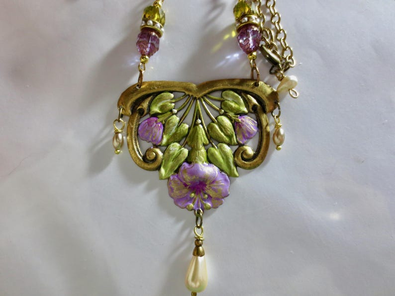 May include: A vintage-style necklace featuring a floral pendant with purple and green painted details. The pendant is suspended from a gold-tone chain and adorned with faux pearls and beads. The necklace has a classic, elegant design.