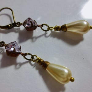 May include: A pair of vintage-style dangle earrings. Each earring features a faceted, light purple crystal, connected by a small chain to a teardrop-shaped faux pearl. The metal components have an antique gold finish.