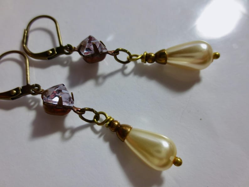 May include: A pair of vintage-style dangle earrings. Each earring features a faceted, light purple crystal, connected by a small chain to a teardrop-shaped faux pearl. The metal components have an antique gold finish.