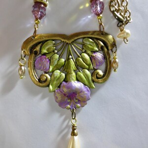 May include: A vintage-style necklace with a floral pendant. The pendant features a gold-toned filigree design with green leaves and purple flowers. The necklace includes a chain, beaded accents, and a pear-shaped pearl drop.