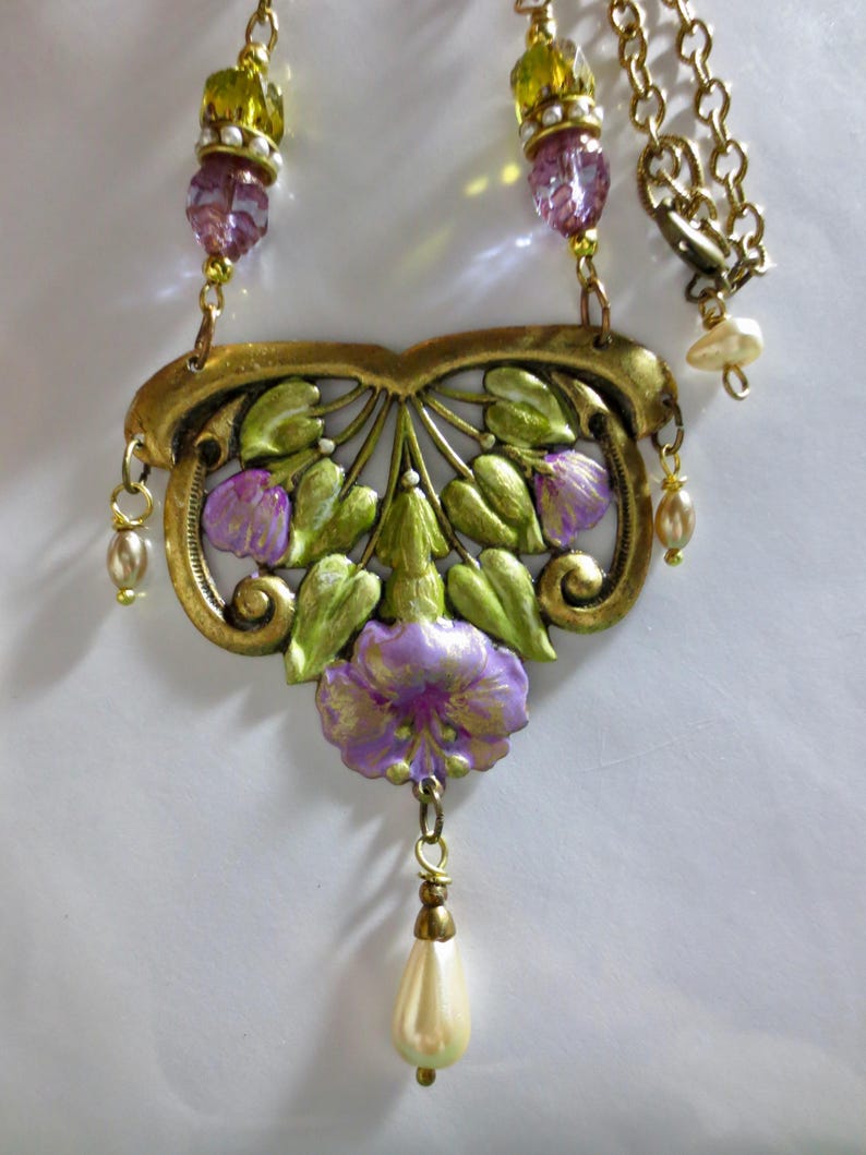May include: A vintage-style necklace with a floral pendant. The pendant features a gold-toned filigree design with green leaves and purple flowers. The necklace includes a chain, beaded accents, and a pear-shaped pearl drop.