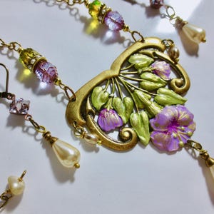 May include: A vintage-style jewelry set featuring a floral pendant necklace and matching earrings. The necklace has a gold-tone filigree pendant with purple flowers and green leaves. The earrings and necklace have pearl and crystal accents.