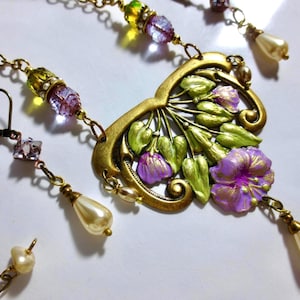 May include: A vintage-style jewelry set featuring a gold-toned necklace and earrings. The necklace has a floral pendant with purple and green accents, complemented by beaded chains and faux pearl drops. The earrings mirror the design with matching beads and pearl drops.