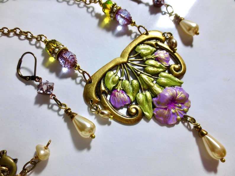 May include: A vintage-style jewelry set featuring a gold-toned necklace and earrings. The necklace has a floral pendant with purple and green accents, complemented by beaded chains and faux pearl drops. The earrings mirror the design with matching beads and pearl drops.