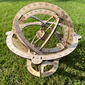 Könnte beinhalten: Eine hölzerne Armillarsphäre mit abgestuften Bögen und beschrifteten Himmelskörpern, darunter Erde und Uranus. Die Sphäre ist aus hellem Holz gefertigt und steht auf einem Sockel auf einer grasbewachsenen Oberfläche.