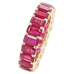 Puede incluir: Un anillo de oro con una fila continua de piedras preciosas rojas rectangulares y vibrantes. La banda del anillo es de color oro cálido, con pequeños detalles dorados entre cada piedra preciosa. Las piedras preciosas son de un color rojo intenso y rico.