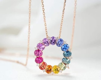May include: A rose gold necklace featuring a circular pendant adorned with a rainbow of oval-cut gemstones. The gemstones include shades of pink, purple, blue, green, yellow, and orange. Small white gemstones are interspersed between the larger stones.