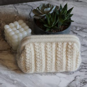 May include: A cream-coloured, cable-knit clutch bag. The rectangular bag has a textured, vertical cable pattern and a smooth, ribbed top. A white bubble candle and potted succulents are in the background, set on a marble surface.