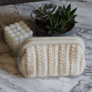 Op de afbeelding: Crèmekleurige clutch van kabelgebreid. De rechthoekige tas heeft een textuur, verticaal kabelpatroon en een gladde, geribbelde bovenkant. Een witte bubbelkaars en vetplanten in pot staan op de achtergrond, op een marmeren oppervlak.