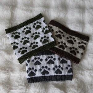 Puede incluir: Tres cuellos tejidos con un diseño de huellas de patas. Cada braga de cuello presenta un fondo blanco con huellas oscuras. Los bordes están ribeteados en verde oscuro, gris oscuro y marrón. Los artículos se muestran sobre una superficie blanca y esponjosa.