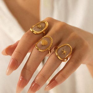 May include: Three gold rings with oval, pale centres. Each ring has a different ocean theme: a sun, a seashell, and a starfish. The rings are displayed on a hand with long, polished nails.