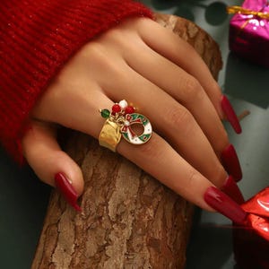 Peut inclure: Une bague dorée ornée d'une couronne de Noël avec des accents rouges, verts et blancs. La bague a une large bande et est ornée de petites perles colorées. La couronne est ornée d'un nœud rouge et de feuilles vertes, parfaite pour les fêtes.