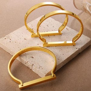 May include: Three gold-coloured, open-ended bangles with a rectangular bar at the base. The bangles are displayed on a textured, light-coloured surface. The jewellery has a polished, reflective finish.
