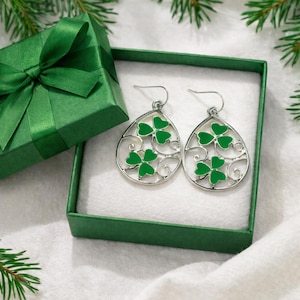 May include: A pair of silver-toned, teardrop-shaped earrings with green shamrock designs, presented in a green gift box with a matching bow. The earrings are set against a white background, with evergreen sprigs in the background.