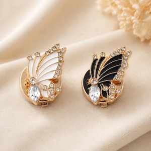 May include: Two butterfly-shaped pins with gold-tone metal frames. One pin features white enamel wings, while the other has black enamel wings. Both pins are embellished with clear round rhinestones along the edges and a single teardrop-shaped crystal.