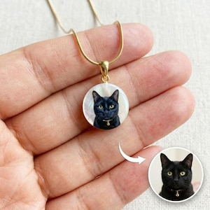 Peut inclure: Un collier doré avec un pendentif rond représentant un portrait de chat noir aux yeux verts. Le chat porte un petit collier avec une clochette. Le pendentif est posé sur un fond blanc.