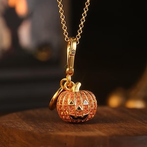 Puede incluir: Un collar dorado con un colgante de calabaza. El dije de calabaza naranja tiene cristales naranjas incrustados, una cara tallada y un pequeño tallo. El colgante cuelga de una cadena dorada, ideal para Halloween.
