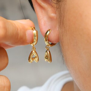 Może przedstawiać: Złote kolczyki koła z wiszącą zawieszką w kształcie litery "A". Kolczyki są pokazane na uchu, jeden z nich jest trzymany przez dłoń. Kolczyki mają błyszczące, wypolerowane wykończenie.