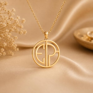 Puede incluir: Un collar de tono dorado con un colgante circular de diseño recortado. El colgante muestra las letras mayúsculas E y P integradas en un marco redondo, suspendido de una delicada cadena de tono dorado a juego con un acabado pulido.